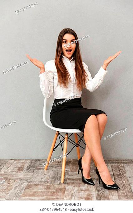 Full length portrait of joyful pretty woman with long brown hair in business wear throwing up hands while sitting on chair isolated over gray background