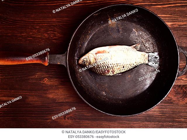 Fresh carp fish in a black cast-iron frying pan on a brown wooden table, top view