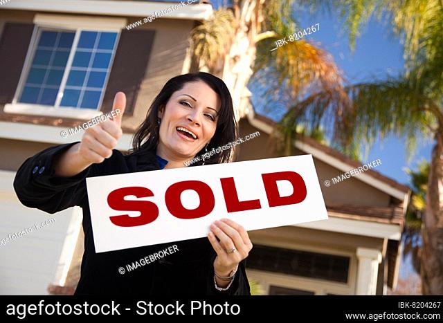 Happy attractive hispanic woman with thumbs up holding sold sign in front of house
