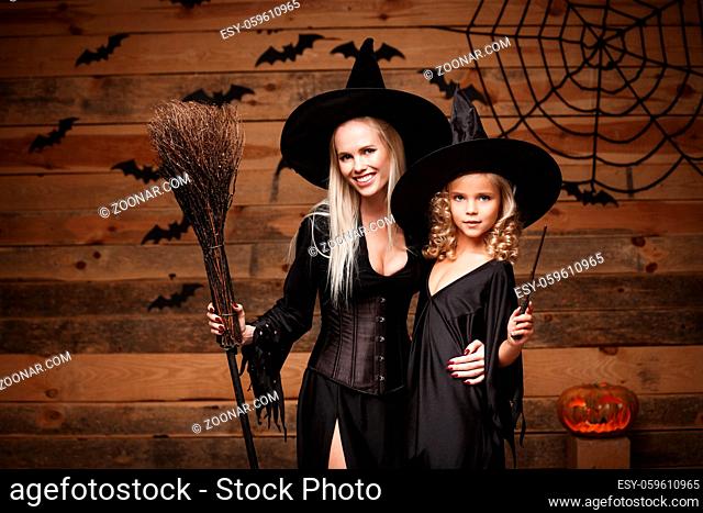 Halloween Concept - cheerful mother and her daughter in witch costumes celebrating Halloween posing with curved pumpkins over bats and spider web on Wooden...