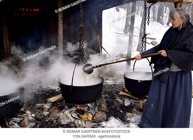 Woman in pioneer clothes ladling evaporated sap in cast iron pots to produce maple syrup