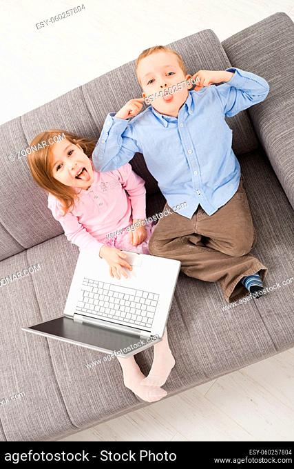 Overhead shot of children sitting on couch at home holding laptop computer, looking up with mocking expression