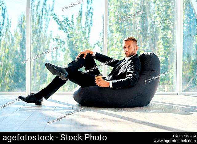 Business man relaxing on bean bag chair with cup of coffee on panoramic windows background in modern creative office with copy space