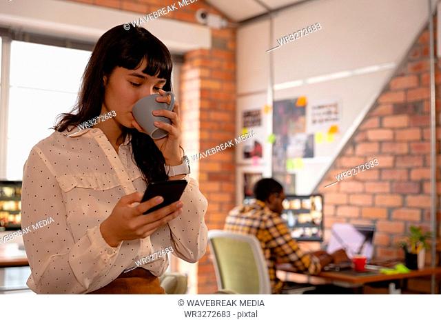 Businesswoman using mobile phone while having cup of coffee in office