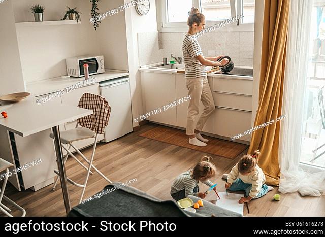 Children drawing together on the sheet of paper with colored pencils while their mother preparing breakfast