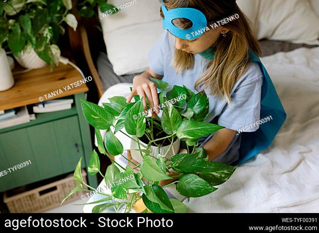 Girl wearing cape and mask sitting with potted plant on bed at home