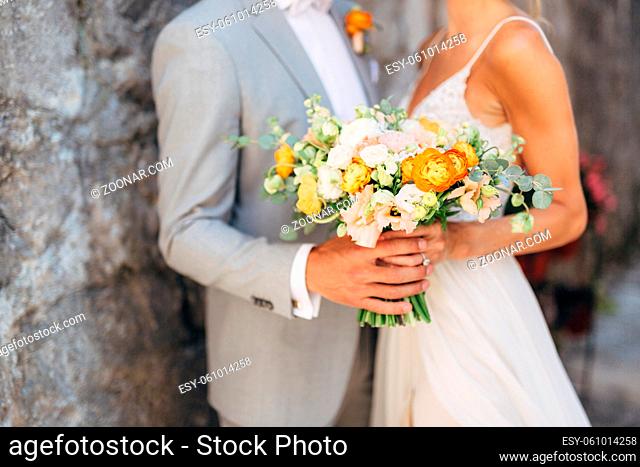 The bride and groom stand hugging at a brick wall in the old city and hold a wedding bouquet with orange flowers, close-up . High quality photo