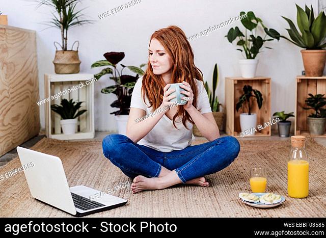 Woman with crossed legs looking at laptop while having coffee on floor at home
