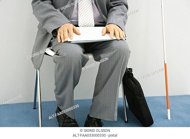Businessman sitting in chair, white cane propped beside him, holding document, cropped