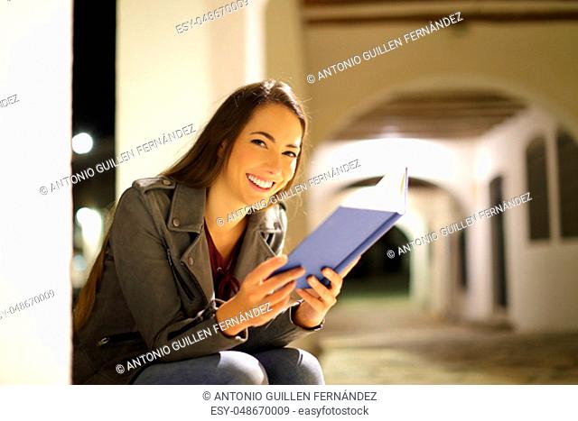 Happy woman looking at camera holding a paper book in the night in the street