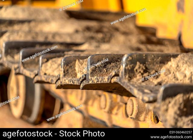 Close up of the metal grouser pad of a yellow contruction machine on a sunny day. The track of the excavator is covered with dry soil