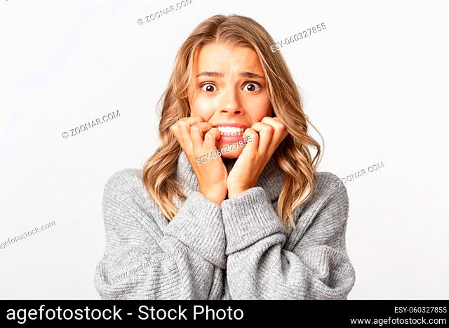 Close-up of scared blond woman in grey sweater, biting fingernails and looking horrified, standing over white background