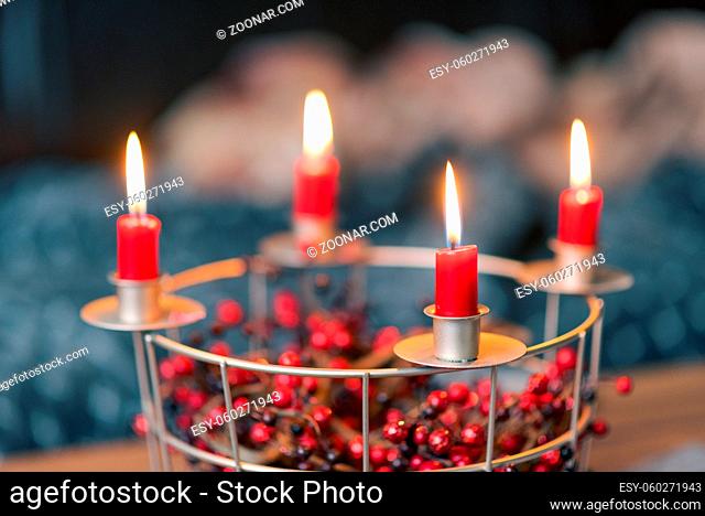 Red candles burning on selfmade Advent wreath made of wire grid decorated with red dried berries with blurred couch living room in background