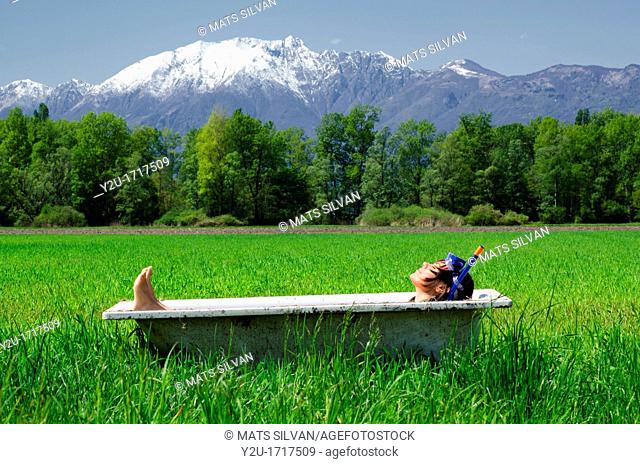 Woman lying in a bathtub on a green field with grass and trees and with snow-capped mountains