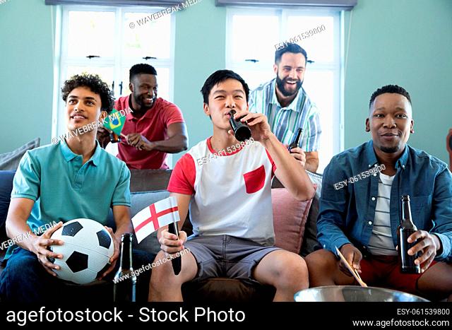 Happy multiracial male friends with ball and beer bottles watching soccer match on sofa at home