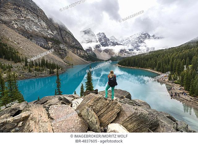 Young woman standing in front of a lake looking into mountain scenery, clouds hanging between mountain peaks, reflection in turquoise lake, Moraine Lake