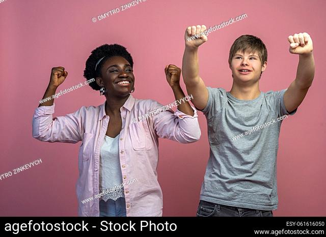 Good mood. Smiling dark-skinned woman looking at caucasian guy with down syndrome standing with raised fists on light background