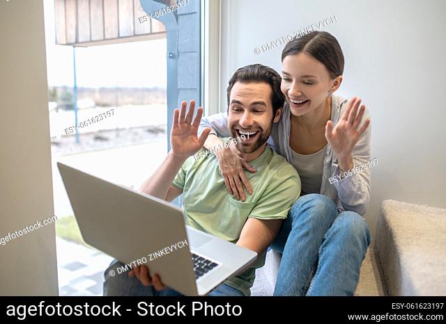 Video call. Smiling young adult man and woman gesturing greeting looking at laptop while sitting near window at home