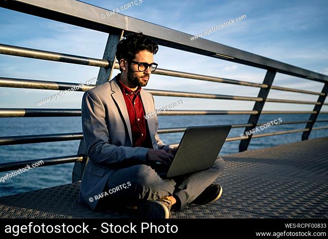 Male business professional using laptop while sitting cross legged on pier