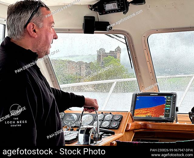 26 August 2023, Great Britain, Drumnadrochit: Skipper Ali Matheson stands on the bridge of a boat on Loch Ness, the ruins of Urquhart Castle can be seen in the...