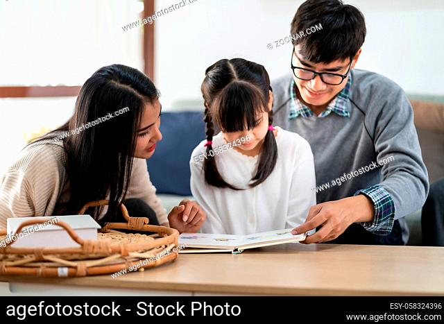 Asian cute girl reading story cartoon book with her mom and dad in living room. Happiness cheerful family and domestic lifestyle concept