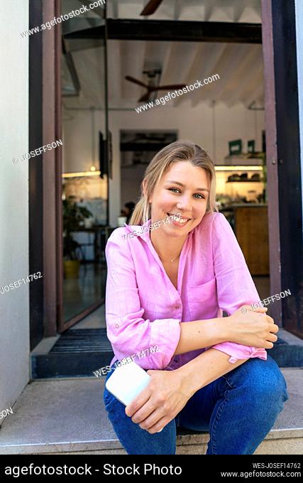 Happy cafe owner sitting with smart phone at doorway