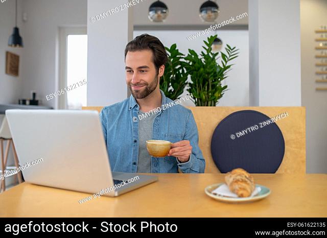 Good mood. Smiling young bearded man with coffee sitting at table with laptop looking at screen in cafe