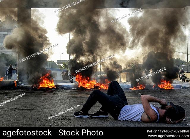 29 November 2021, Lebanon, Beirut: A man lies on the ground in front of burning tires blocking a main highway in Beirut during a protest against the dire...