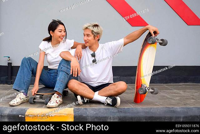 Asian couple resting after skateboarding. A young man in a white T-shirt held his hand on the surfskates board while talking to his girlfriend