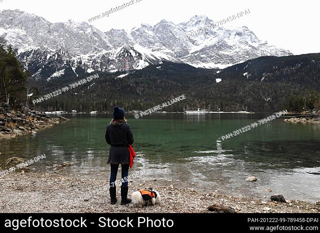 22 December 2020, Bavaria, Eibsee: A hiker stands with her dog at the Eibsee in front of the backdrop of the Zugspitze. With mild temperatures and slightly...