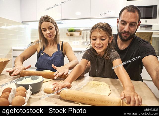 Parents with smiling daughter kneading pizza dough on table in kitchen