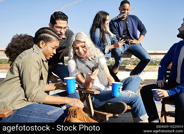 Group of young co-workers hanging out on rooftop patio laughing and having a drink, sharing images on mobile devices