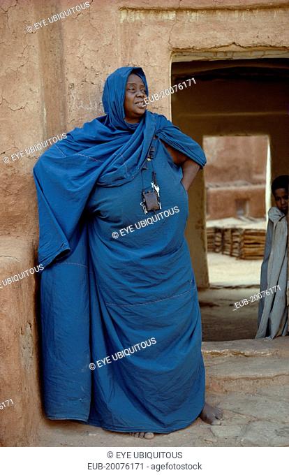 Portrait of woman wearing turquoise robe and head covering standing in front of mud brick building with child framed in doorway behind her