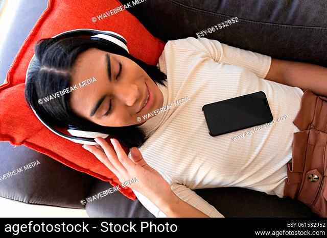 Smiling mixed race gender fluid man lying on couch wearing wireless headphones using smartphone