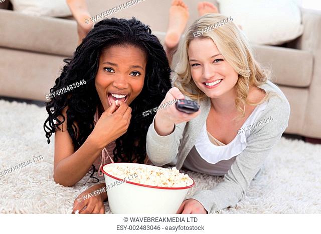 Two women lying on the ground with a bowl of popcorn and a TV romote