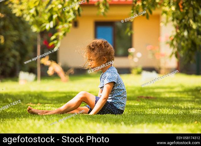Side view of a little blonde girl sitting on a green lawn. The girl is sitting with her legs outstretched in the backyard