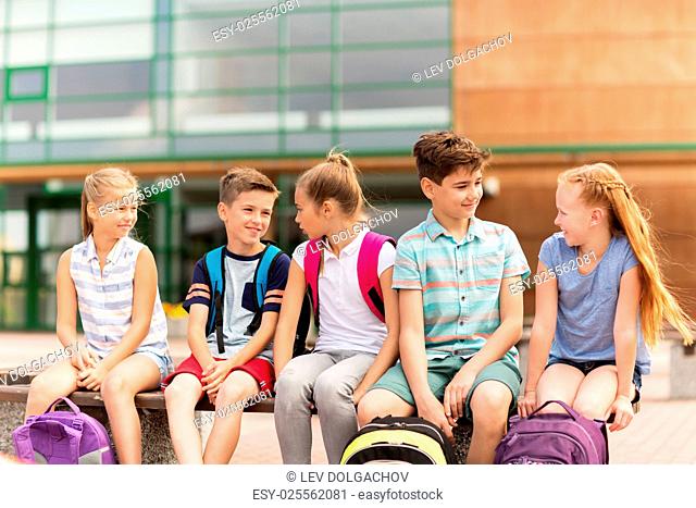 primary education, friendship, childhood, communication and people concept - group of happy elementary school students with backpacks sitting on bench and...