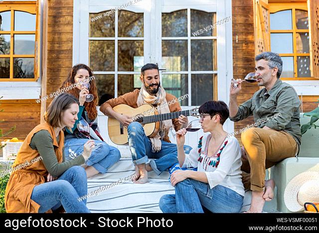 Friends playing music on the guitar and drinking wine outside a cabin in the countryside