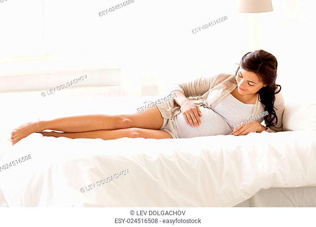 pregnancy, rest, people and expectation concept - happy pregnant woman lying in bed and touching her belly at home bedroom