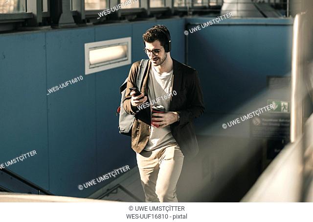 Smiling young man with headphones, cell phone and takeaway coffee walking at the station