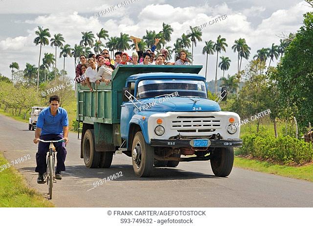 A dump truck full of people passing a man riding a bicycle