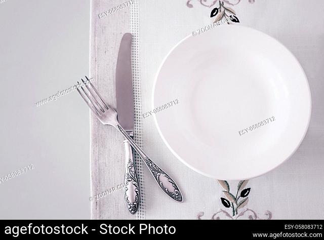 On the bright surface of the table is a white empty plate on a linen napkin . Next to a knife and fork. Top view, copy space