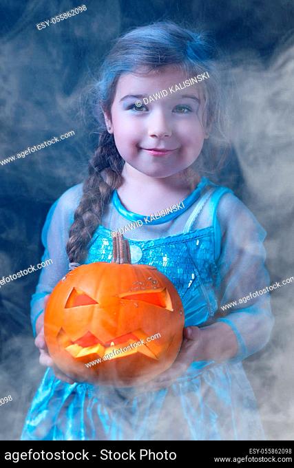 Halloween portrait of young girl stylised for frozen princess in a blue dress. Smiling child holding orange curved pumpkin surrounded by smoke or mist