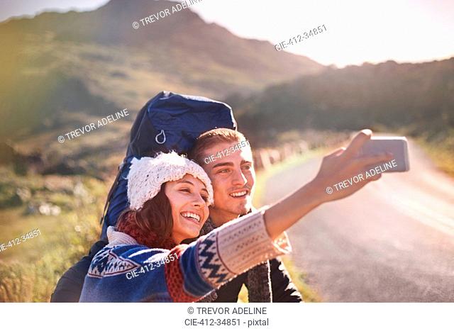 Young couple with backpacks hiking taking selfie with camera phone on sunny, remote road