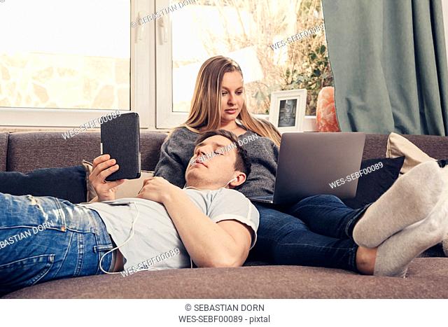 Young couple relaxing on sofa and using notebook, smartphone and ebook reader