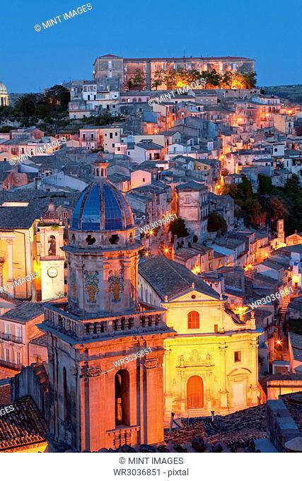 High angle view over illuminated rooftops of traditional houses in a Mediterranean city at dusk, church cupola in the foreground