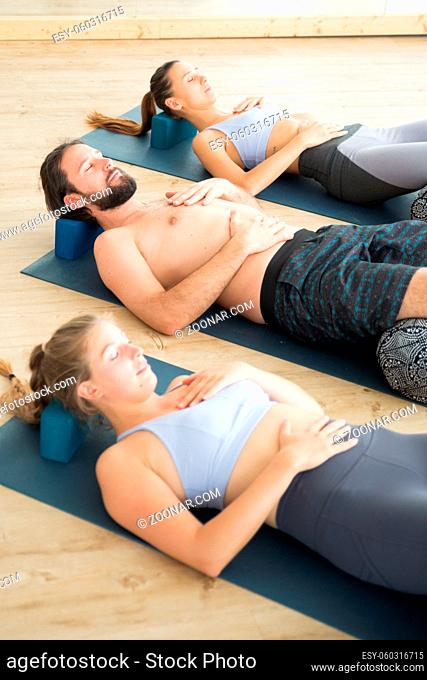 Restorative yoga with a bolster. Group of young sporty attractive people in yoga studio, lying on bolster cushion, stretching and relaxing during restorative...