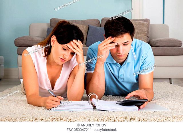 Young Unhappy Couple Calculating Bills While Lying On Carpet At Home