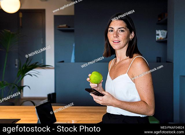 Smiling young woman with apple and mobile phone at home