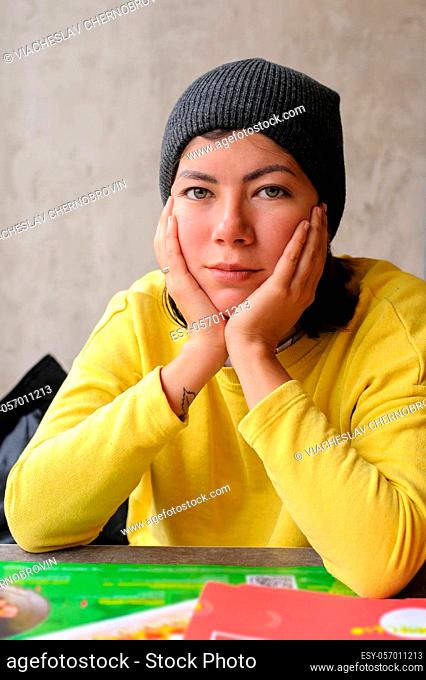 a cute young woman in a knit hat and a yellow sweater sits at a table in a cafe, resting her head on two folded hands